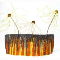 A 3D fine woven tapestry in orange, yellow and grey wools, with circular woven yellow and orange fishing line flowers sitting above the tapestry, their wire stems embedded in the woollen weave 