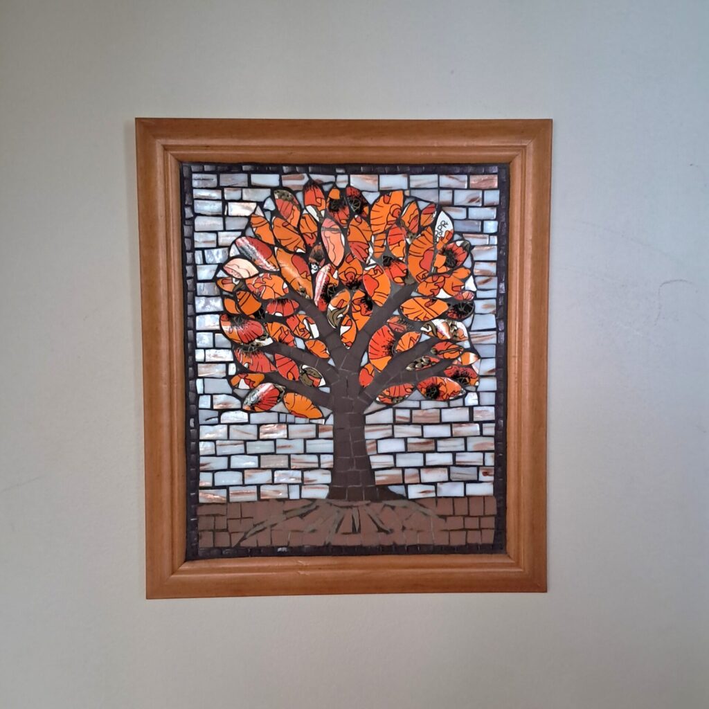Framed 'Flame Tree' mosaic with vibrant orange & black leaves made from 'Meakin Poppy' ceramics. The trunk provides contrast in both colour & material, being of unglazed dark brown ceramic. The background of gilded white glass suggests evening & helps to highlight the tree.