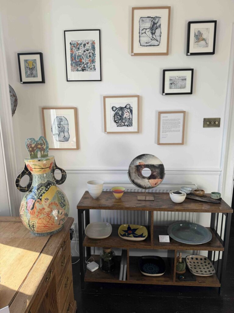 A bright gallery-style display in a Georgian townhouse interior. A white wall holds a salon-style arrangement of framed artworks, including birds, a tiger, and a colourful Bishop’s Stortford print. Below, a dark wood shelving unit displays ceramic pieces: bowls, platters, small vessels, and a circular sculptural platter on a stand. To the left, a large hand-painted ceramic vase with bold, playful decoration sits on a wooden chest of drawers. Sunlight falls across the room, giving the space a warm, curated feel.